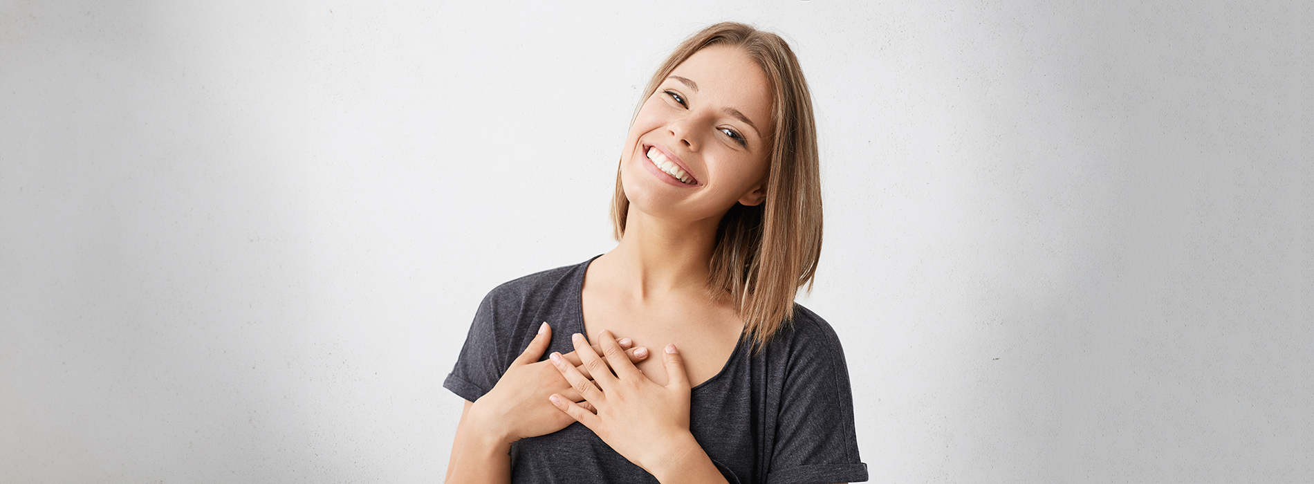 The image is a photograph of a woman with light skin, smiling at the camera. She appears to be in her late twenties or early thirties and has long hair. Her eyes are looking directly at the camera, and she is holding up her index finger near her mouth as if she s making a point or emphasizing something. The background is plain and light-colored, which suggests that this could be a stock photo used for various purposes such as advertising, personal branding, or lifestyle content.